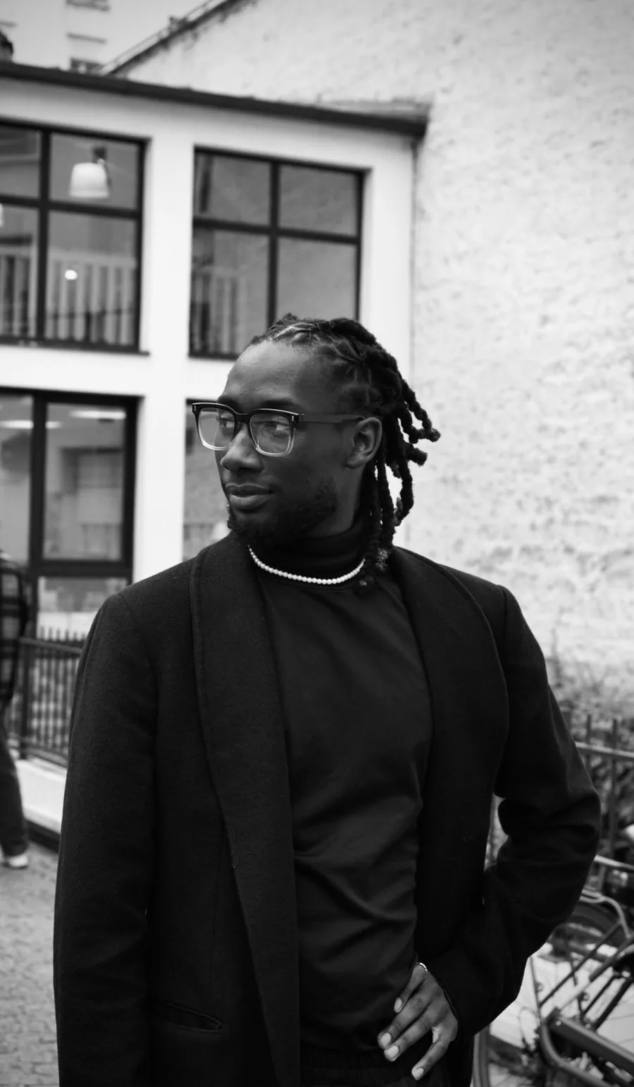 Portrait en noir et blanc de Djibril Sacko, manteau noir et col roulé, devant une façade parisienne.